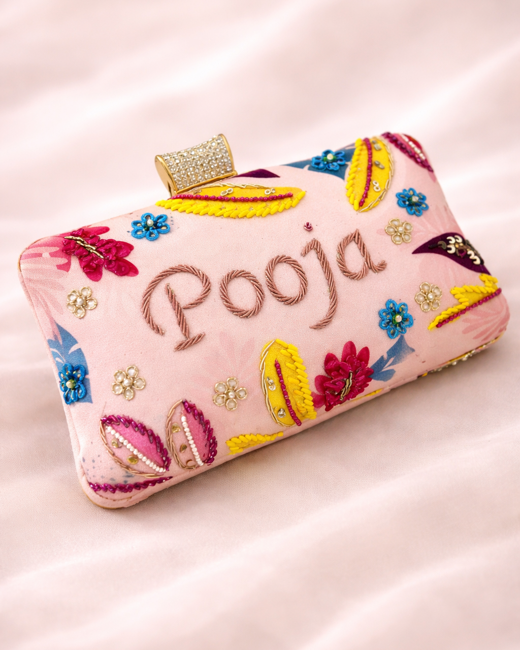 Colorful clutch bag with floral designs and 'Pooja' embroidery on a pink background